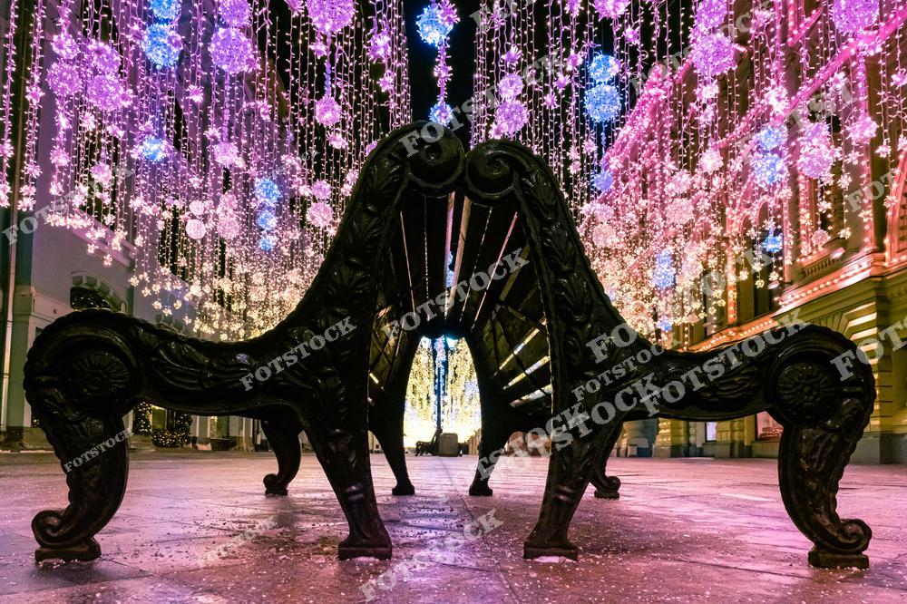 Фотография демонстрирует декоративные скамейки, расположенные на пешеходной улице в Москве, украшенной праздничной иллюминацией. Разноцветные гирлянды и световые шары, выполненные в розовых, голубых и золотистых тонах, создают ощущение волшебства и праздника. Окружающие здания освещены мягкими огнями, подчеркивая архитектурные детали. Центральное расположение скамейки акцентирует внимание на уютной атмосфере улицы. Эта зимняя сцена отражает красоту и романтику столицы в период новогодних праздников.